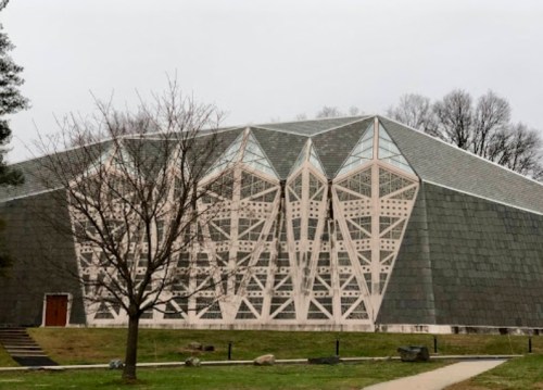 A large, modern building with a geometric facade, surrounded by trees and a grassy area on a cloudy day.