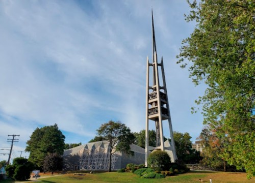 A tall, modern church tower with a spire, surrounded by trees and a grassy area under a clear blue sky.