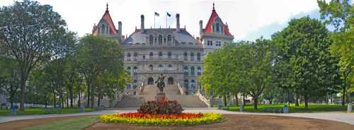 Panoramic view of a grand building with red-roofed towers, surrounded by trees and colorful flower beds.