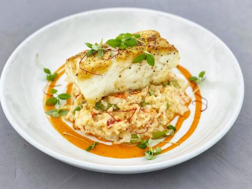 A plate of risotto topped with a piece of seared fish, garnished with microgreens and drizzled with sauce.