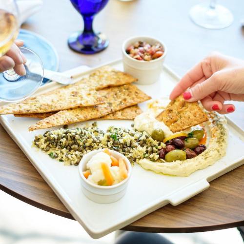 A hand dips a cracker into hummus, surrounded by olives, grains, and a small bowl of salsa on a wooden table.