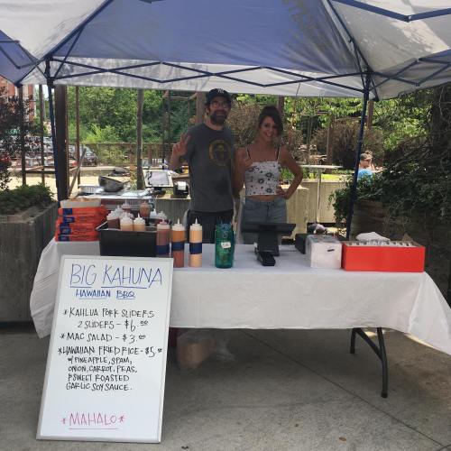 A food stall with two vendors, offering Hawaiian BBQ items like sliders and mac salad, under a blue tent.