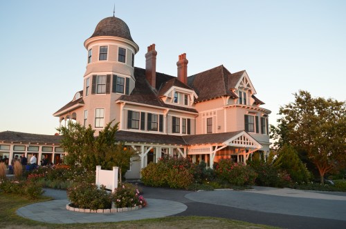 A large, elegant Victorian-style house with a turret, surrounded by gardens and a circular driveway at sunset.