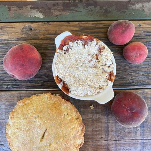 A rustic wooden table with a peach cobbler in a dish, surrounded by fresh peaches.