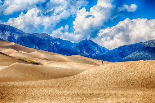 A vast desert landscape with rolling sand dunes and mountains under a blue sky with fluffy clouds.