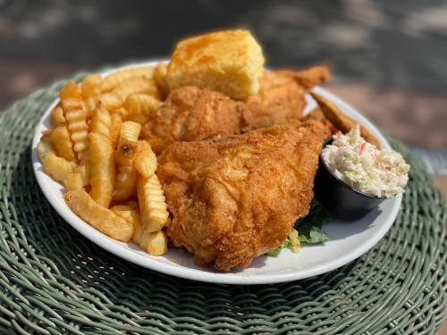 A plate of fried chicken with crinkle-cut fries, coleslaw, and a biscuit, served on a green woven table.