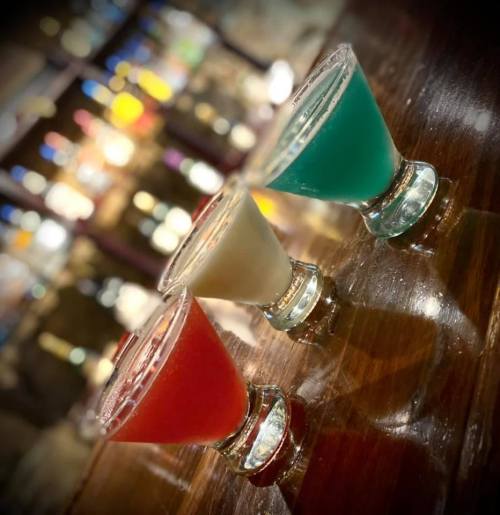 Three colorful cocktails in glass cups on a wooden bar, with a blurred background of bottles.