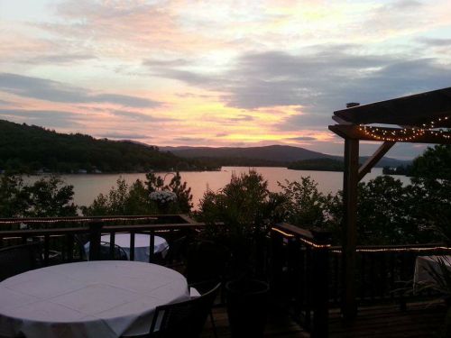 A serene lakeside view at sunset, with mountains in the background and a deck adorned with lights.