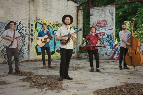 A band of four musicians poses in an urban setting with graffiti, holding various instruments like a banjo and guitar.