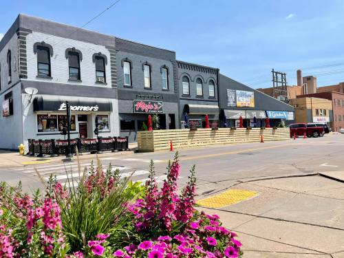 A street view of a block with various shops and restaurants, featuring outdoor seating and colorful flowers in the foreground.