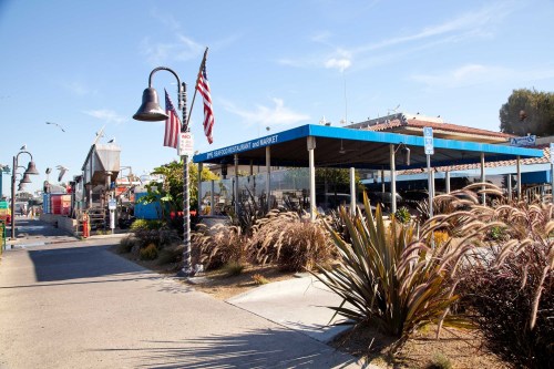 A seaside restaurant and market with outdoor seating, surrounded by plants and an American flag, under a clear blue sky.