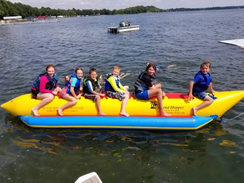 A group of children in life jackets enjoying a banana boat ride on a lake, with a sunny sky in the background.