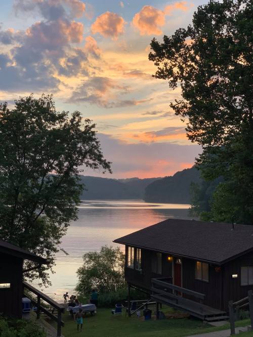 A serene lakeside scene at sunset, with colorful clouds reflecting on the water and a cabin in the foreground.