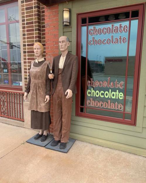 Two life-sized statues of a man and woman in vintage clothing stand outside a chocolate shop, with a colorful sign behind them.