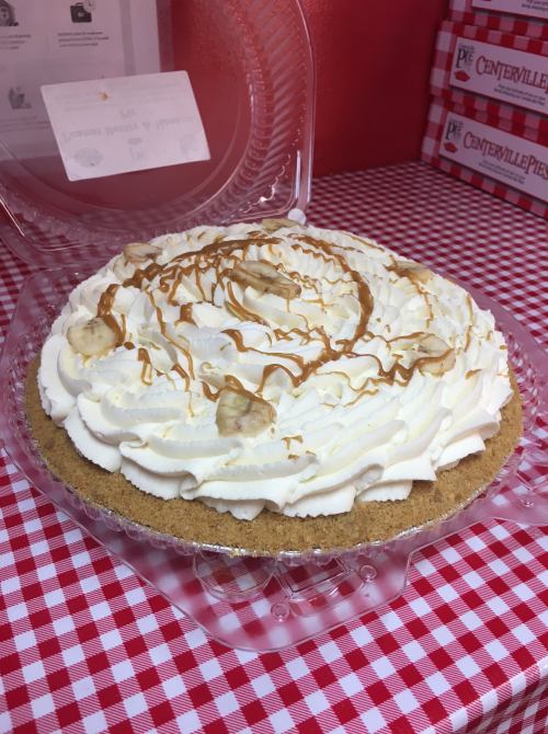 A creamy banana cream pie topped with whipped cream and caramel drizzle, displayed in a clear plastic container.