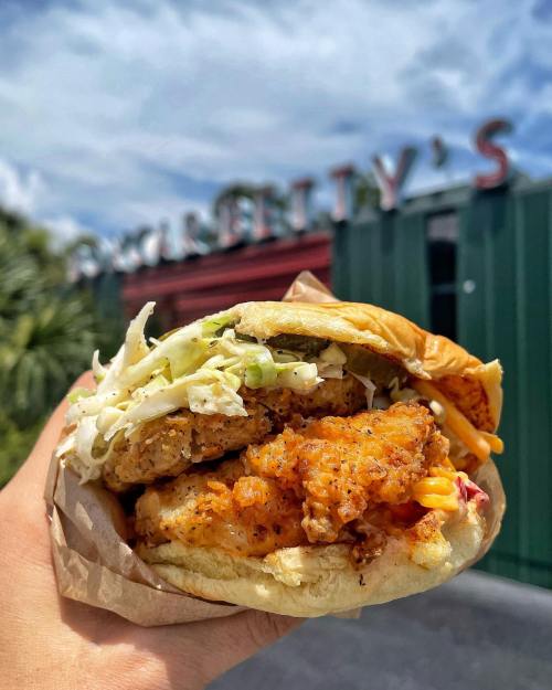A hand holding a crispy chicken sandwich topped with lettuce, against a blurred background of a restaurant sign.