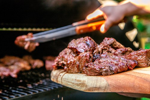 A hand using tongs to place grilled meat onto a wooden platter, with a barbecue grill in the background.