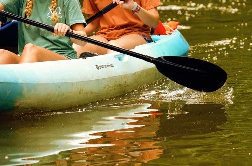 Two people kayaking on a calm water surface, with paddles creating ripples in the water.