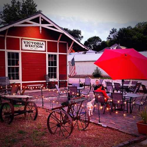 A cozy outdoor seating area with string lights, a red umbrella, and a historic red building labeled "Victoria Station."