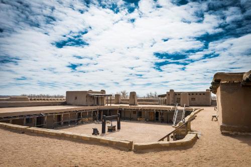 Desert landscape featuring adobe structures under a cloudy blue sky, with a central courtyard and sandy ground.