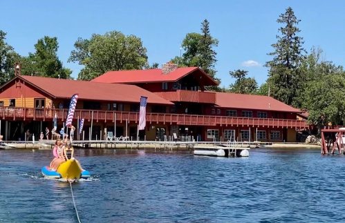 A group of people on a banana boat being pulled near a lakeside lodge with American flags and trees in the background.