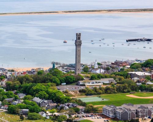 Aerial view of a coastal town featuring a tall tower, sandy beach, and boats in the water. Lush greenery surrounds the area.