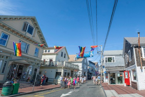A vibrant street scene with rainbow flags hanging above, featuring shops and people walking in a sunny coastal town.
