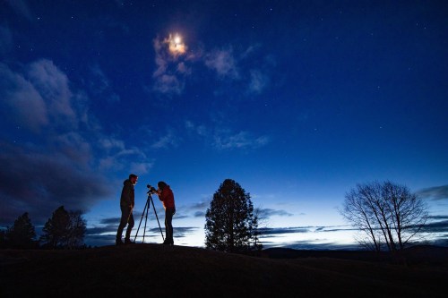 Two photographers set up tripods under a starry sky, with a bright moon and clouds in the background.