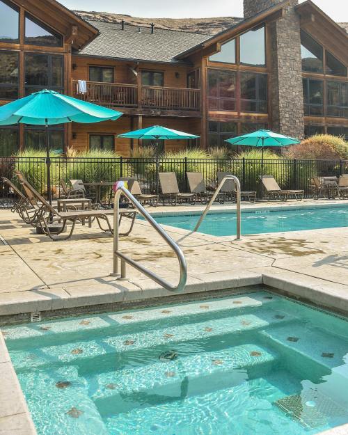A pool area with a hot tub, lounge chairs, and umbrellas, set against a wooden building and mountains in the background.