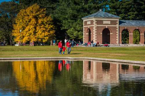 A park scene with people walking by a pond, surrounded by trees with autumn colors and a brick structure in the background.