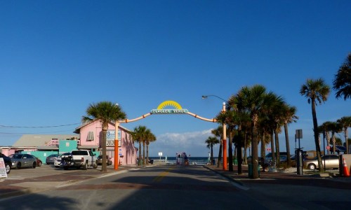 Colorful entrance archway to a beach area, flanked by palm trees and buildings, under a clear blue sky.