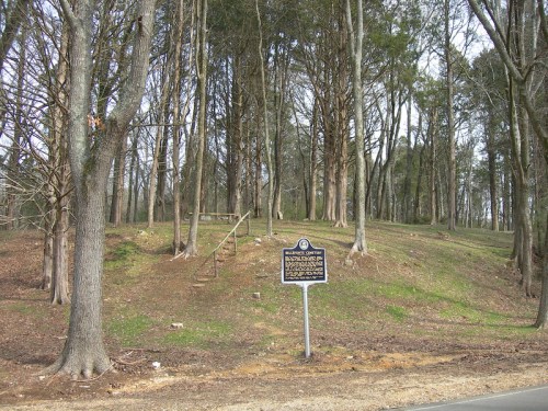 A wooded area with trees and a historical marker sign in the foreground, indicating a significant site.