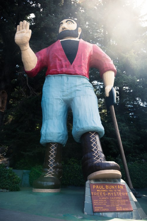 A giant statue of Paul Bunyan, a lumberjack, stands tall among trees, waving with a blue sky above.