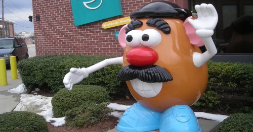A large, cartoonish Mr. Potato Head statue with a mustache and blue shoes, waving outside a building.