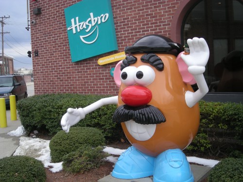 A large Mr. Potato Head statue waves outside the Hasbro headquarters, surrounded by greenery and snow.