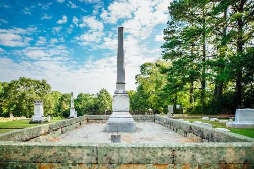 A tall gravestone stands in a serene cemetery surrounded by trees and a clear blue sky.