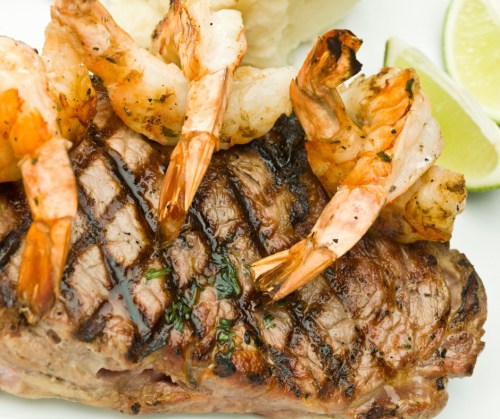 Grilled steak topped with shrimp, served with lime wedges and a side of mashed potatoes.