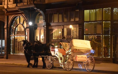 A horse-drawn carriage waits outside a restaurant, illuminated by festive lights at night.
