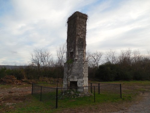 A weathered stone chimney covered in vines, surrounded by a black fence, set against a cloudy sky and bare trees.