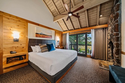 Cozy hotel room with a king bed, wooden accents, and a balcony overlooking greenery. Stone fireplace adds warmth.