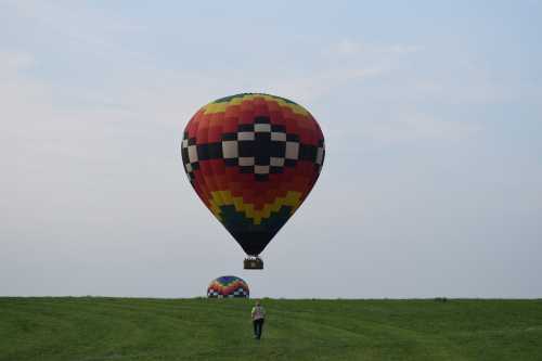 A colorful hot air balloon floats above a grassy field, with a person walking towards it in the foreground.