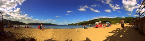 A panoramic view of a sandy beach with people enjoying the sun, water, and colorful umbrellas under a blue sky.