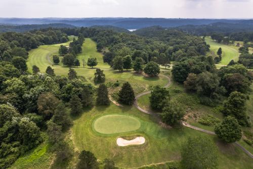 Aerial view of a lush golf course surrounded by trees and rolling hills under a cloudy sky.