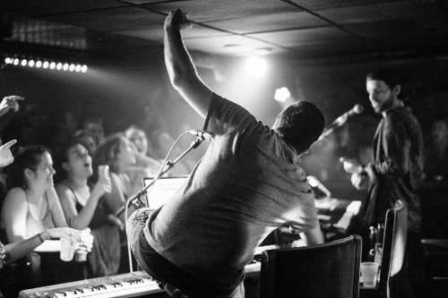 A musician performs on stage while an enthusiastic crowd cheers, captured in a lively black-and-white setting.