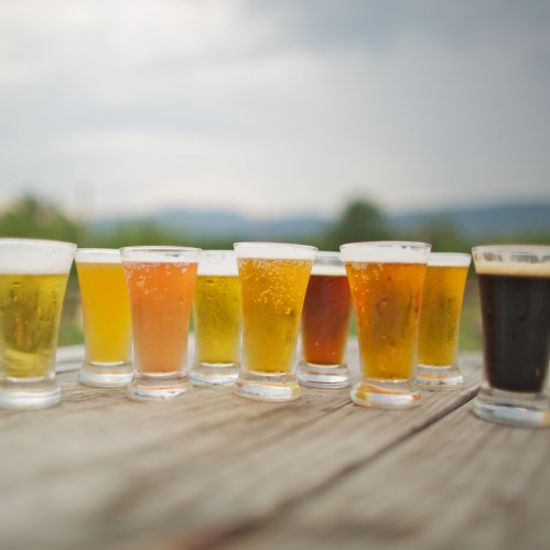 A row of various beer glasses in different colors, set on a wooden table with a scenic background.