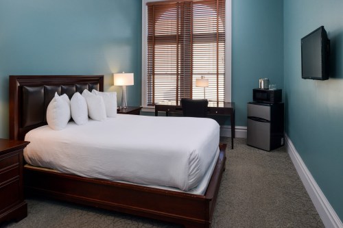 Cozy bedroom with a queen bed, desk, TV, and mini fridge, featuring teal walls and wooden blinds.
