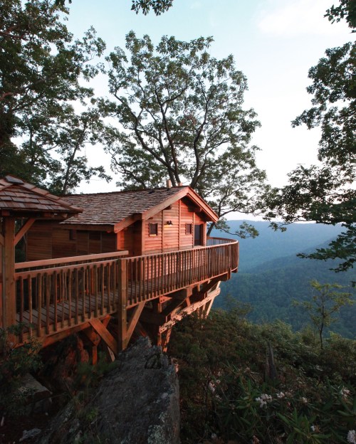 A wooden cabin perched on a cliff, surrounded by trees, overlooking a scenic mountain landscape at sunset.