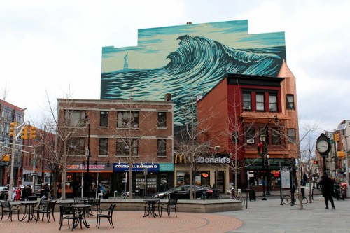 A large mural of ocean waves on a building, with shops and a clock tower in the foreground.