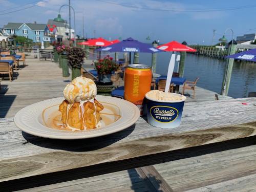 A plate of ice cream with toppings and a cup of ice cream on a wooden table by the water, with colorful umbrellas in the background.