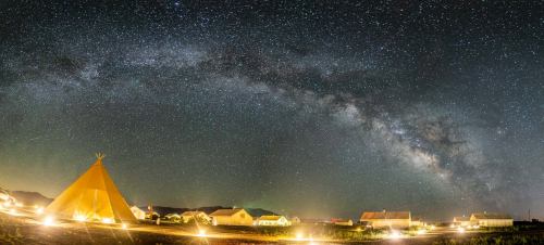 A panoramic view of a starry night sky with the Milky Way over illuminated tents and cabins in a serene landscape.